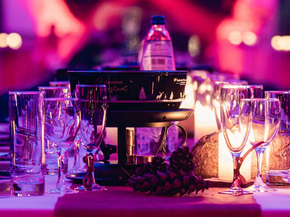 Table de banquet dressée avec un caquelon à fondue, des verres et des pommes de pin, sous des lumières roses et violettes